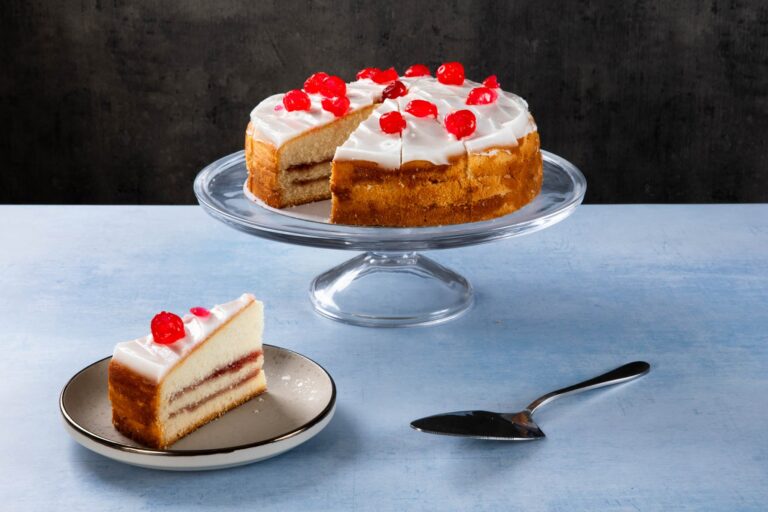 Cherry-topped sponge cake with white frosting on a glass cake stand.