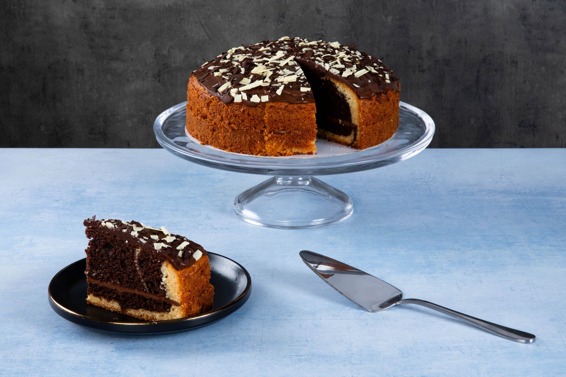 Chocolate cake with chocolate topping and white chocolate shavings on a glass cake stand.
