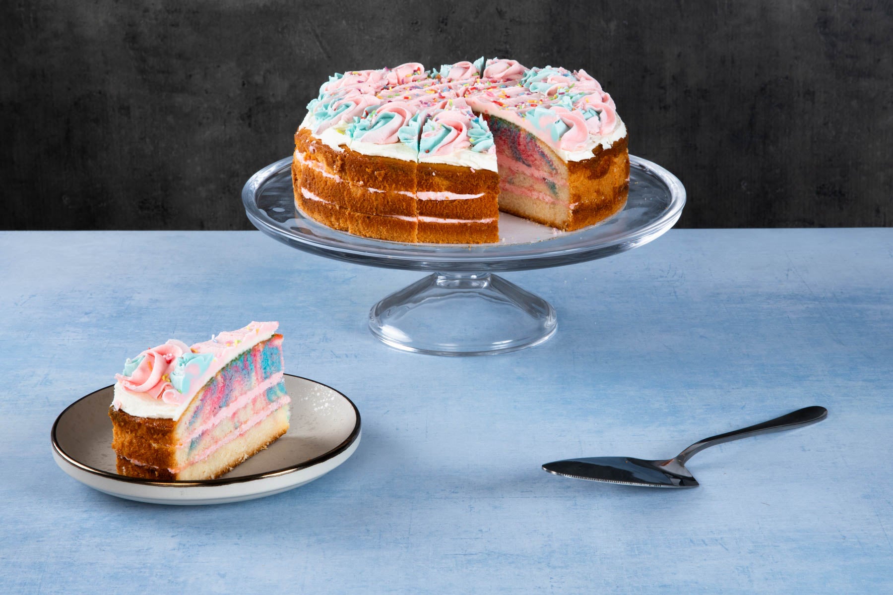 Rainbow cake with colourful layers and white frosting on a glass cake stand.
