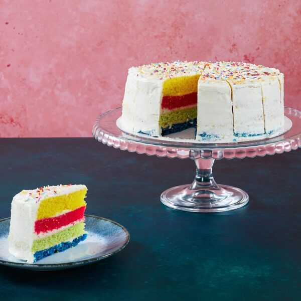 Rainbow cake with colourful sponge layers and sprinkles on a glass cake stand.