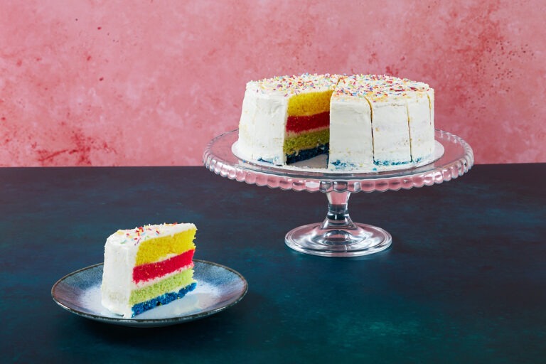 Rainbow cake with colourful sponge layers and sprinkles on a glass cake stand.