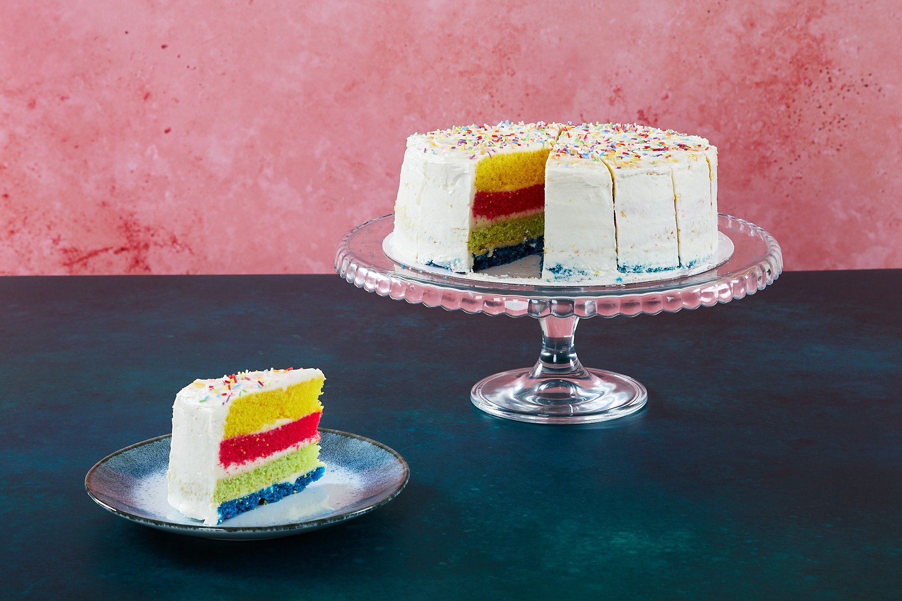 Rainbow cake with colourful sponge layers and sprinkles on a glass cake stand.