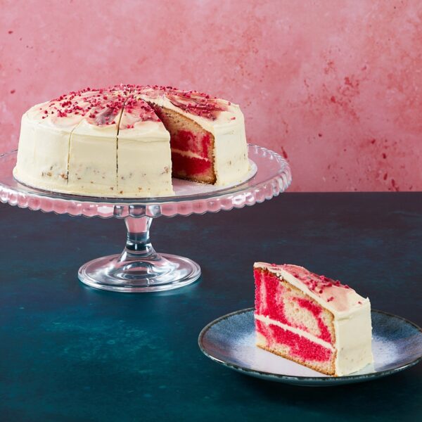 Raspberry ripple cake with raspberry frosting and topping on a glass cake stand.