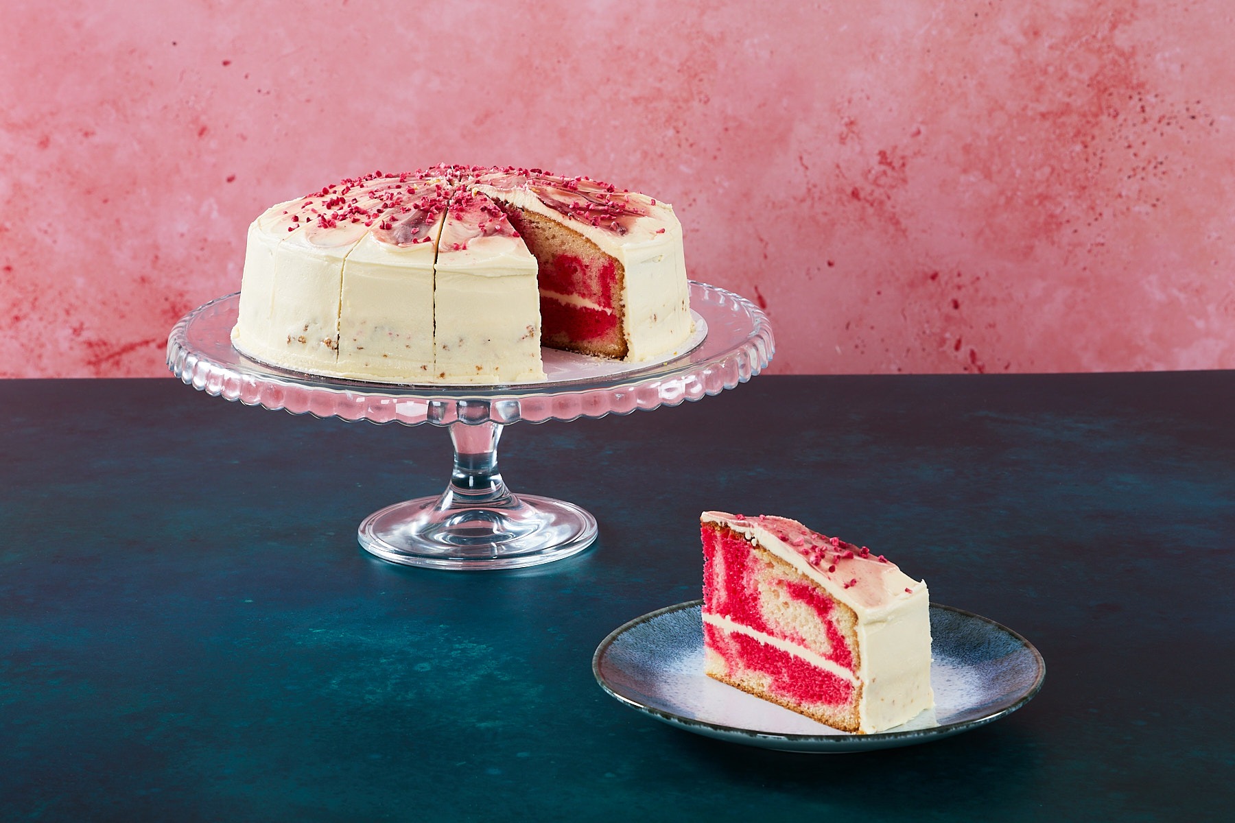 Raspberry ripple cake with raspberry frosting and topping on a glass cake stand.